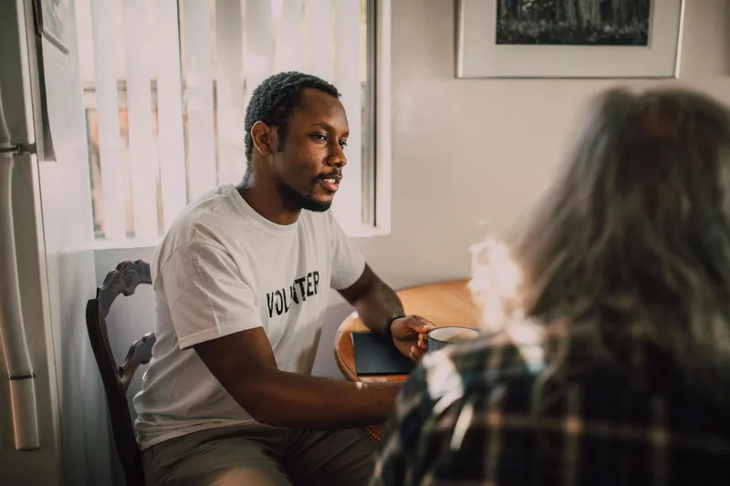 Two people in a coffee shop having a focused conversation, one listening and taking notes